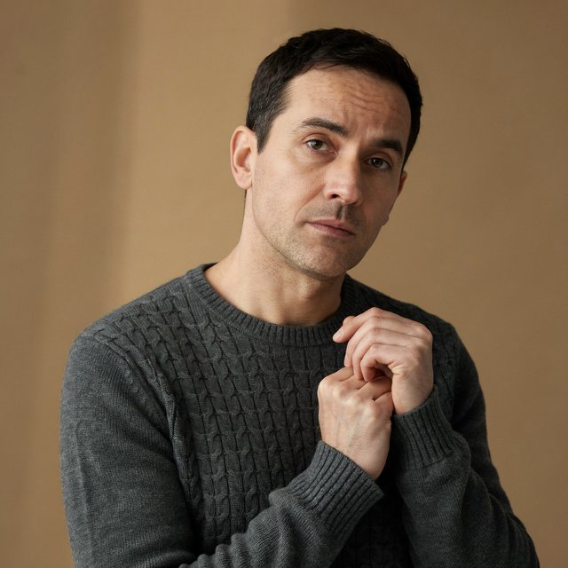 Editorial portrait of an adult man in his early 40s wearing a charcoal wool fisherman’s sweater, one hand loosely holding its ribbed cuff. His thoughtful, slightly tired expression rests against a muted ochre studio backdrop, shaped by gentle directional window light. — Ideogram v4.5