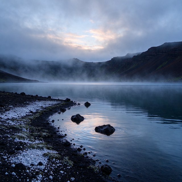 At first light, a rain-cleared volcanic caldera lake lies beneath thinning fog; pumice grains and damp lichen sharpen the near shore, while dark cinder slopes step back toward a distant rim. — Seedream 5.0 Flash