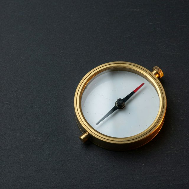 A compact brass pocket compass rests on black slate, its satin-metal casing and clear glass cover catching a broad diffused key and narrow warm rim light against a deep charcoal background; an unbranded commercial product image, blank face without lettering. — Grok Imagine Image Quality