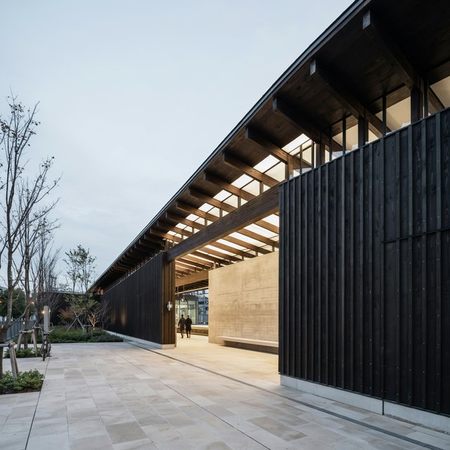 Contemporary Japanese timber train station, charred cedar cladding, pale concrete platform, broad pitched roof with exposed beams and translucent clerestories, cool morning light, viewed obliquely from the quiet approach plaza, precise straight lines and plausible structure. — Ideogram v4