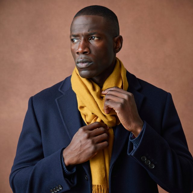 Editorial portrait of an adult Black man in his 30s wearing a deep navy wool overcoat and mustard scarf, fingers adjusting the scarf, thoughtful expression, muted terracotta studio backdrop, crisp cool window light. — ImagineArt 2