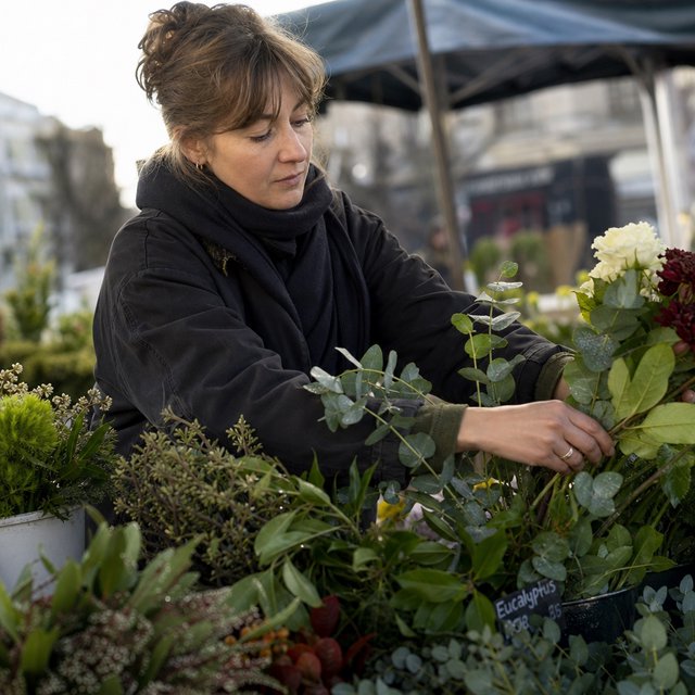 Candid documentary photograph of an adult florist arranging loose stems at an outdoor market stall, soft early-morning side light, 50mm lens with shallow depth of field, tiny water droplets beading on the cut leaves. — GPT Image 2
