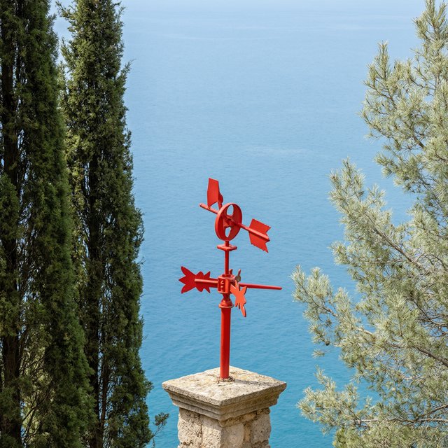 At a coastal overlook, a small red weather vane stands on a stone post. Exactly two dark-green pine trees line the left edge and one pale-green pine tree stands at the right edge; a blue sea fills the background, nothing else. — Model 18