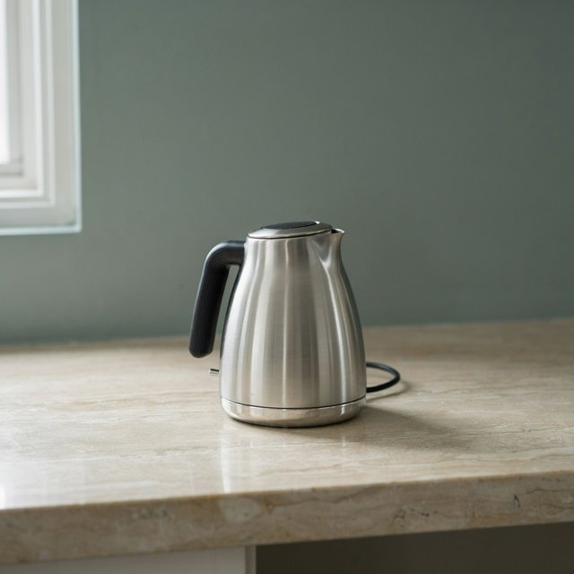 Morning daylight spills across a kitchen counter, revealing an unbranded electric kettle in satin stainless steel with a ribbed heat-resistant polymer handle. It sits on honed limestone against a muted sage backsplash, with window light and white bounce fill; commercial product image, no lettering. — Ideogram v4.5