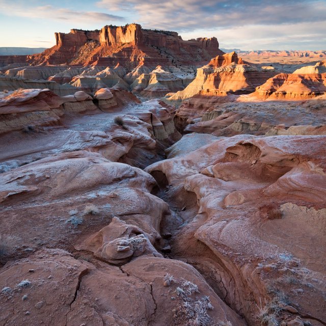 A red-sandstone badlands plateau beneath clearing winter clouds at sunrise, overnight frost silvering the gullies; cracked clay and wind-smoothed rock lead the eye across layered mesas fading into pale distance. — ImagineArt 2