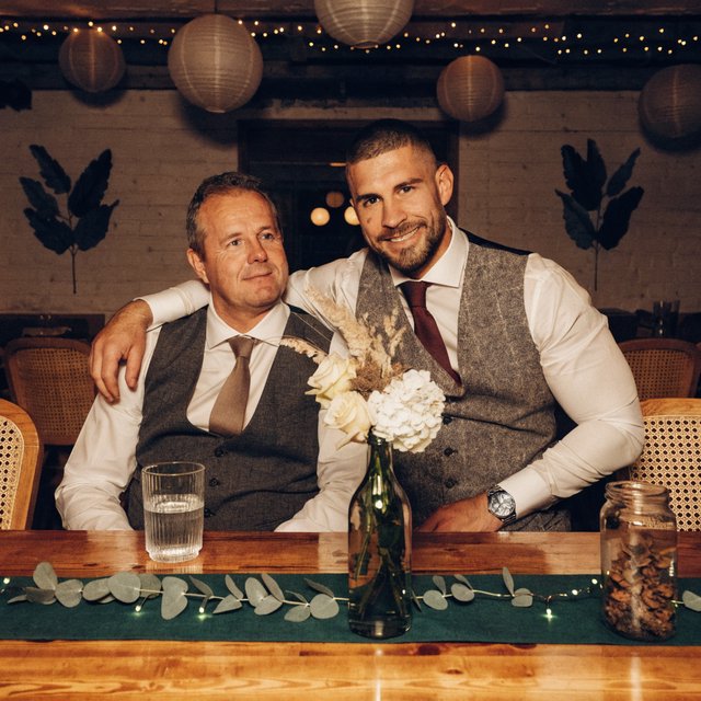 (Character1: rugby player body type, tall, brunet male, age 30, buzzcut hair and neatly groomed matching beard). Medium eye-level shot, photorealistic, two men seated at a wooden banquet table: the left middle-aged man wears a white dress shirt, charcoal waistcoat, and taupe tie, with his right arm on the table; the right man, Character1, smiles directly at the camera, wearing a white dress shirt, grey waistcoat, maroon tie, and a watch on his left wrist, with his left arm draped over the first man's shoulder. In the foreground: a table runner with eucalyptus sprigs, small fairy lights, a bottle holding flowers, a glass jar, and a water glass. Background: an exposed light horizontal wood plank wall with two decorative leaf panels, an exposed industrial ceiling with hanging paper lanterns, and fairy string lights. Warm, soft ambient overhead event lighting, natural indoor venue atmosphere, shallow depth of field. Art style: dramatic painterly realism with realistic skin texture, strong chiaroscuro, slightly gritty textured brushwork, fantasy illustration mood. — Recraft v4
