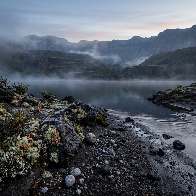 At first light, a rain-cleared volcanic caldera lake lies beneath thinning fog; pumice grains and damp lichen sharpen the near shore, while dark cinder slopes step back toward a distant rim. — Nano Banana 2.1
