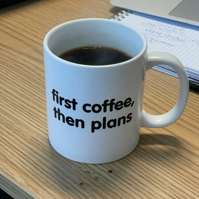 A white ceramic mug on a desk with the words "first coffee, then plans" printed in black lowercase sans-serif. — Grok Imagine Image Quality