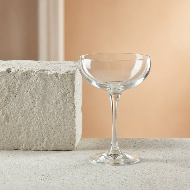 Beside a pale limestone block, an unbranded handblown cocktail coupe in clear soda-lime glass catches crisp window-shaped highlights; its curved bowl bends a muted apricot backdrop under broad diffused key light and narrow cool rim lighting, commercial product image, no lettering. — Grok Imagine Image 2