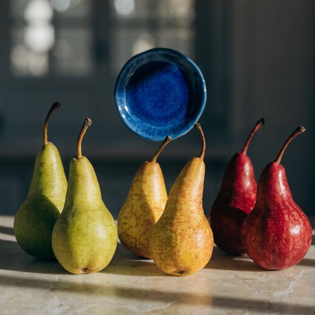 Exactly six ripe pears sit on a pale stone counter: two green pears on the left, two yellow pears in the center, and two red pears on the right. A small blue plate sits above the center pair, nothing else. — Recraft v4