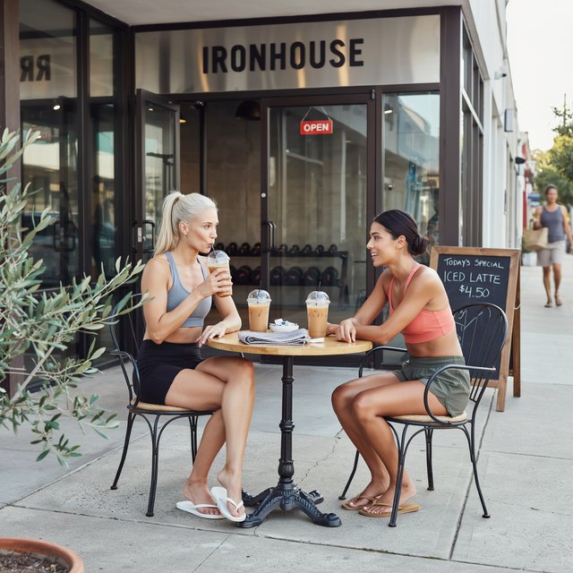 Two muscular adult women in their twenties, one Caucasian blonde and one Latina, sit at an outdoor café table beside a gym after their workout. They wear tight workout shorts, sports bras, and flip-flops. — Ideogram v4