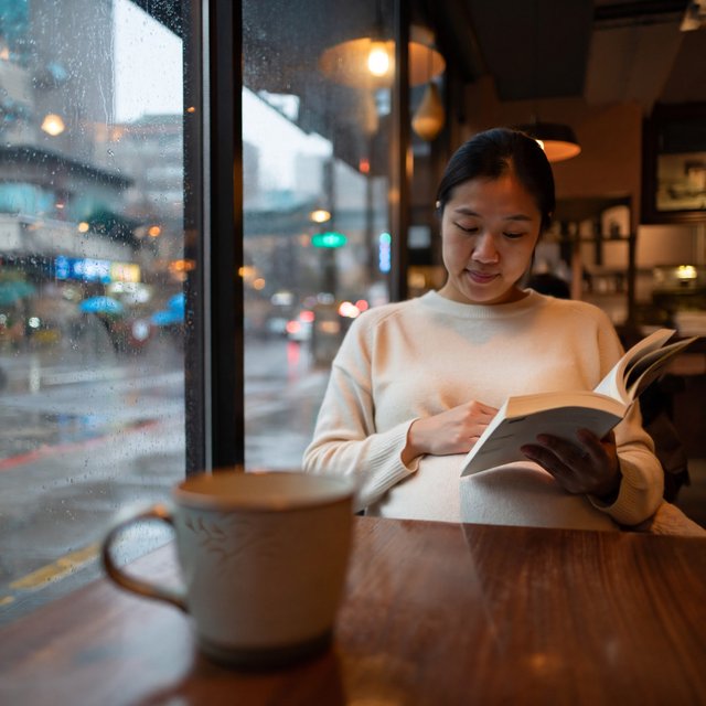 Photorealistic cinematic scene of a pregnant Chinese woman sitting in a cozy café by the window, reading a book. She appears calm, gentle, and thoughtful, with one hand resting naturally near her belly. On the table beside her is a cup of coffee, softly steaming. Outside the large window, heavy rain pours down, with streaks of water on the glass and a blurred rainy street beyond. The café interior is warm and inviting, with soft ambient lighting, wooden textures, and a peaceful atmosphere that contrasts with the storm outside. The composition should feel intimate, emotional, and serene, with the woman as the clear focal point. — ImagineArt 2