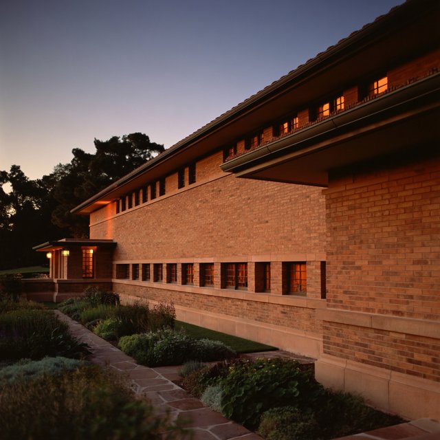Exterior of a Prairie School residence with broad overhanging eaves, warm brick, limestone courses, and ribbon windows; amber evening light grazes the facade, viewed obliquely from a garden path, with disciplined horizontal lines and credible structure. — ImagineArt 2