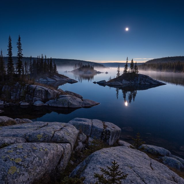 An expansive boreal lake beneath a clear, cold midnight sky, pale moonlight catching thin mist above dark water; spruce-covered islands recede toward distant ridges, while lichen and cracked granite sharpen the near shoreline. — Recraft v4