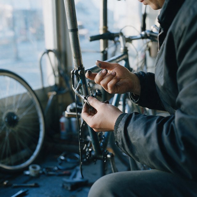 Candid documentary photograph of an adult repairing a bicycle at a quiet curbside workshop, cool morning window light, 50mm lens with shallow depth of field, grease smudges on their fingertips. — Seedream 5.0 Pro
