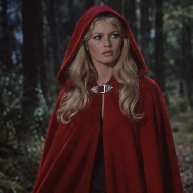 Medium shot at eye level: 1960s vintage film aesthetic. An adult woman with long, blonde hair walks alone through a dark forest. She wears a red hooded cloak, with the hood pulled up and fastened at the neck by a silver buckle. Photorealistic 1960s Technicolor film grain; soft, shallow depth of field. — GPT Image 2.5 Flare