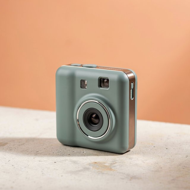 Commercial product photograph of an unbranded compact instant camera in matte forest-green polymer with brushed steel details, resting on a pale concrete surface against a warm peach background, illuminated by broad diffused studio light and a soft rim light. — Ideogram v4.5