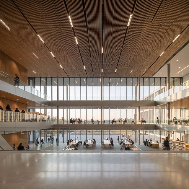 Interior of a modern public library with a three-storey atrium, timber ceiling slats, people reading, afternoon light through a glass wall. — ImagineArt 2