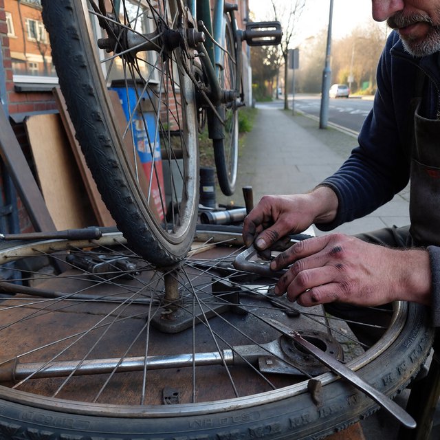 Candid documentary photograph of an adult repairing a bicycle at a quiet curbside workshop, cool morning window light, 50mm lens with shallow depth of field, grease smudges on their fingertips. — ImagineArt 1.5 Pro