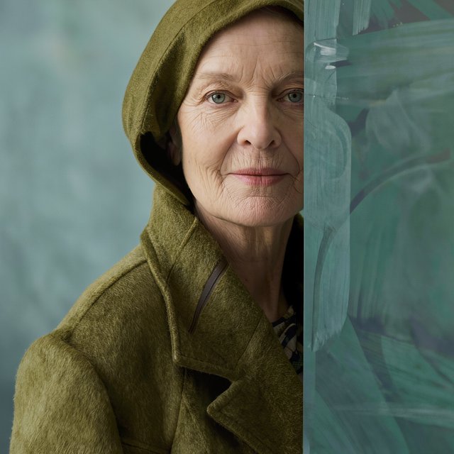 Beside a faded teal studio wall, a close-up fashion portrait frames an adult woman aged 68–76, her weathered skin softly detailed; she wears a moss-green wool coat and holds a calm, searching gaze in gentle side light. — Model 9