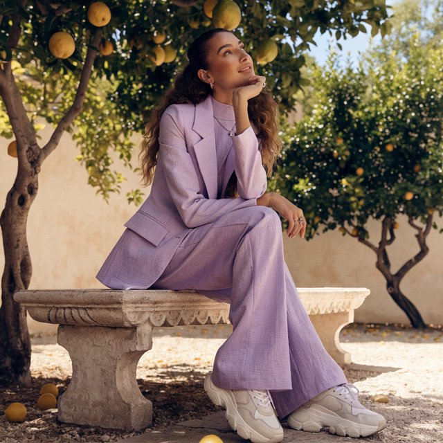 Editorial glamour photograph of one adult woman in a tailored lilac fitness set, seated naturally on a sun-warmed stone bench in a quiet citrus courtyard, flattering dappled light, loose curls, thoughtful smile, crisp leaves and pale stucco. — Recraft v4