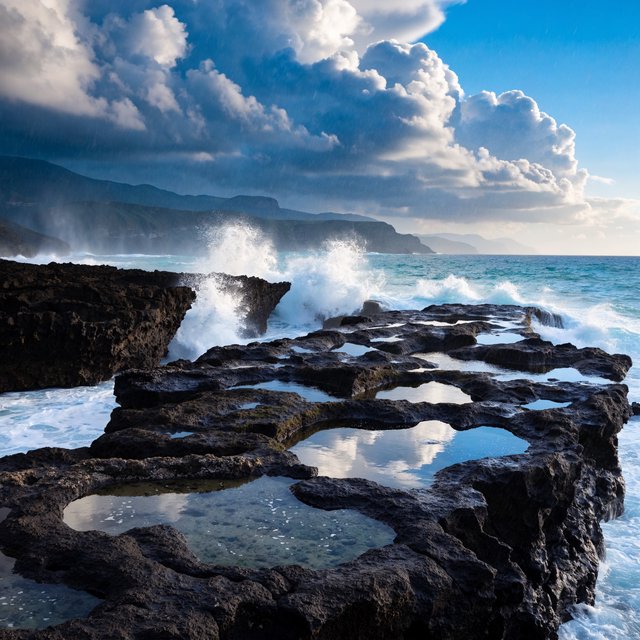 A rugged volcanic coastline under clearing storm clouds at late afternoon, black basalt shelves descending into turquoise surf, sea spray clinging to jagged rocks, tide pools reflecting distant cliffs and layered mountains fading into rain haze. — Recraft v4 Pro
