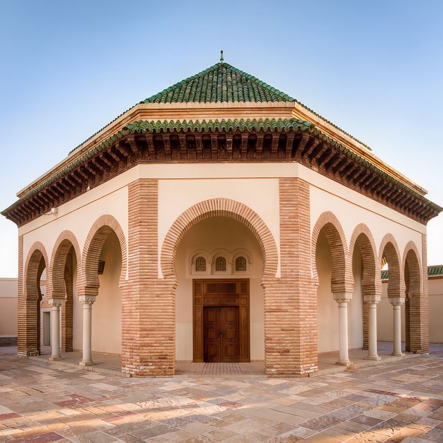 Exterior of a Moorish Revival municipal bathhouse, pale buff stucco, striped brick arches, green glazed tile roof and carved cedar doors; clear morning light, viewed across a paved courtyard, with coherent structure and crisp lines. — Kling v3