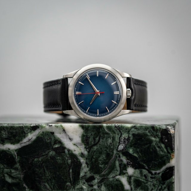 Commercial product photograph of an unbranded mechanical wristwatch with a brushed titanium case and deep blue enamel dial, resting on dark green marble against a soft gray background, illuminated by a large diffused softbox and narrow edge light. — Ideogram v4.5