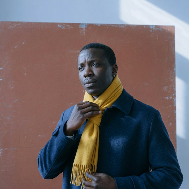 Editorial portrait of an adult Black man in his 30s wearing a deep navy wool overcoat and mustard scarf, fingers adjusting the scarf, thoughtful expression, muted terracotta studio backdrop, crisp cool window light. — Grok Imagine Image Quality