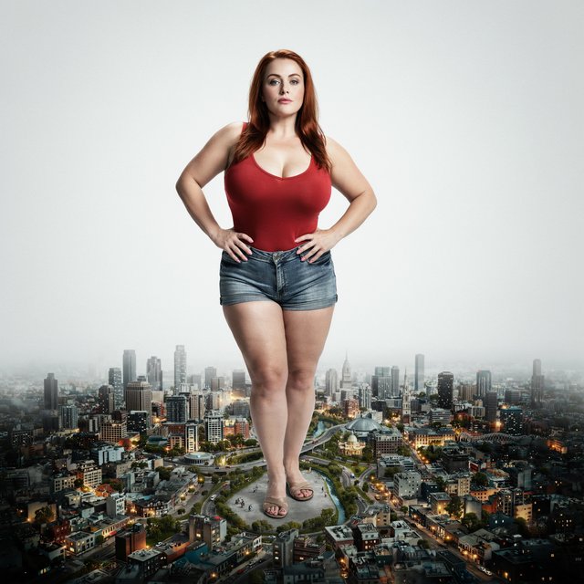 A beautiful adult woman with red hair, pale skin, and blue eyes. She has a slim but curvy body, a very large bust, and thick thighs. She has long pink manicured nails and is wearing a red low-cut singlet and tight denim shorts, along with light makeup. She is standing in a large empty space. A realistic-looking miniature city covers the whole floor, and the woman is standing in the middle of it with her hands on her hips, looking at the viewer. — Ideogram v4
