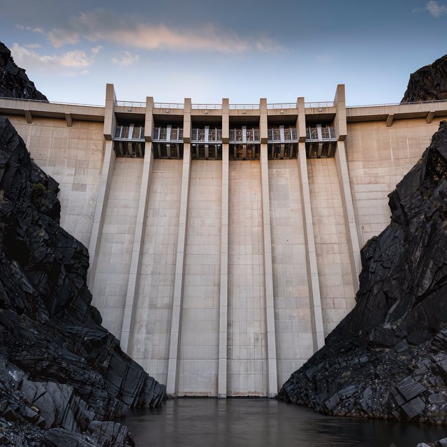 A broad hydroelectric dam in contemporary Brutalist engineering style spans a rocky gorge, its pale concrete face anchored by dark basalt abutments and steel gates; cool dawn light, viewed from downstream, with convincing load-bearing geometry. — Model 9
