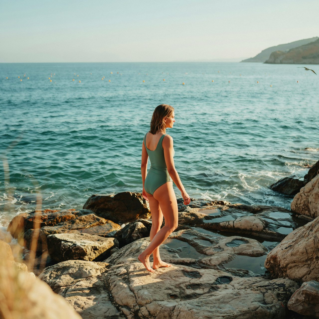 Golden late-afternoon light skims a sheltered cove as an adult woman in a jade one-piece swimsuit is caught mid-step across sunlit rocks, her gaze turned toward the sea; crisp reflections and composed styling bring fashion-magazine polish. — Ideogram v4