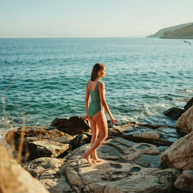 Golden late-afternoon light skims a sheltered cove as an adult woman in a jade one-piece swimsuit is caught mid-step across sunlit rocks, her gaze turned toward the sea; crisp reflections and composed styling bring fashion-magazine polish. — Ideogram v4