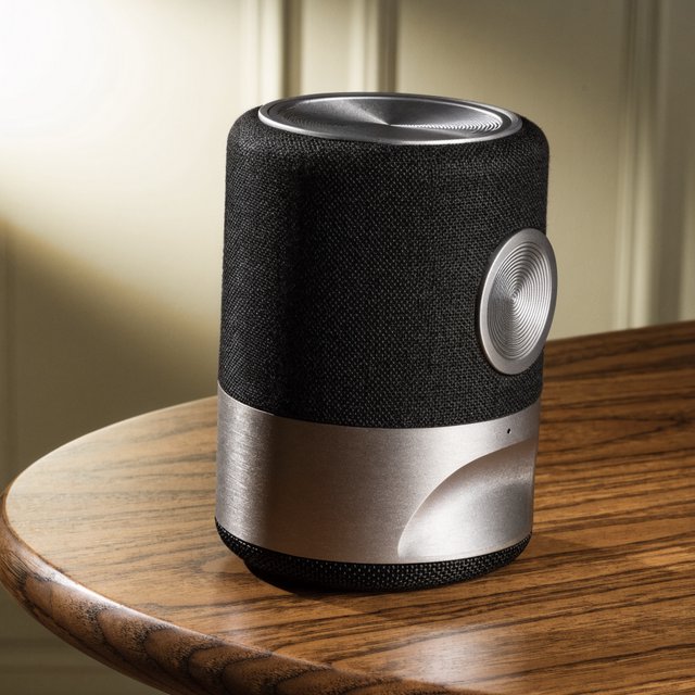 Commercial product photograph of an unbranded wireless speaker made from textured black fabric and satin aluminum, placed on a walnut tabletop against a warm cream background, illuminated by a large softbox and subtle backlight. — Recraft v4