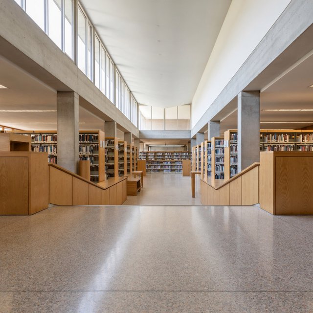 Interior of a mid-century modernist public library, with pale oak shelving, exposed concrete columns, terrazzo flooring, and a broad clerestory. Diffuse morning light crosses the reading hall, viewed from the entrance down its central aisle, with precise lines and plausible structure. — ImagineArt 2