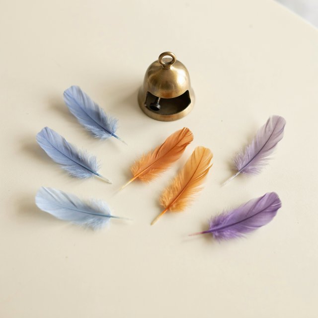 Exactly seven feathers lie on a cream tabletop: three blue on the left, two orange in the center, and two purple on the right. A brass bell sits above the orange pair, nothing else. — Ideogram v4.5