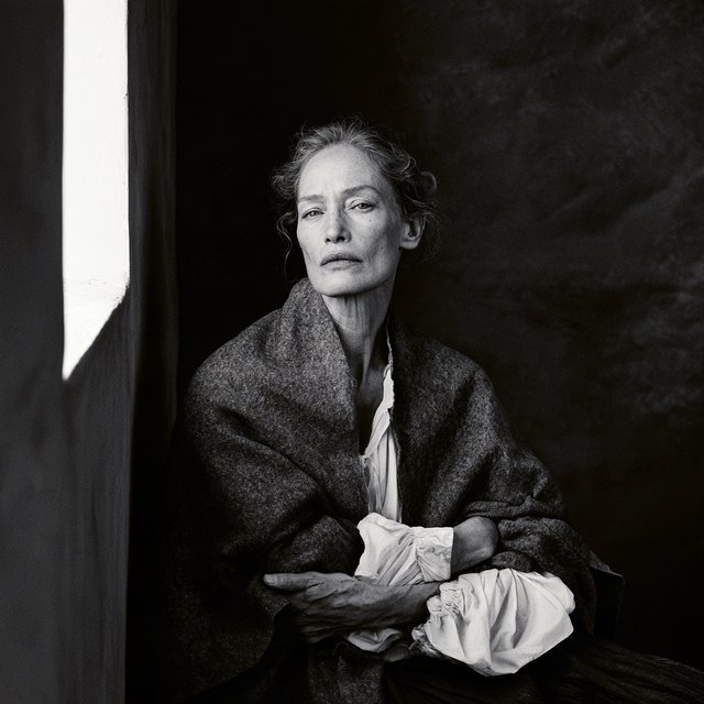 Under a narrow window beam, an adult woman in her late 50s wears a sculpted wool cape over a cotton blouse; her wry expression rests against a charcoal plaster backdrop in a black-and-white portrait, shaped by soft angled light. — Midjourney V8.2