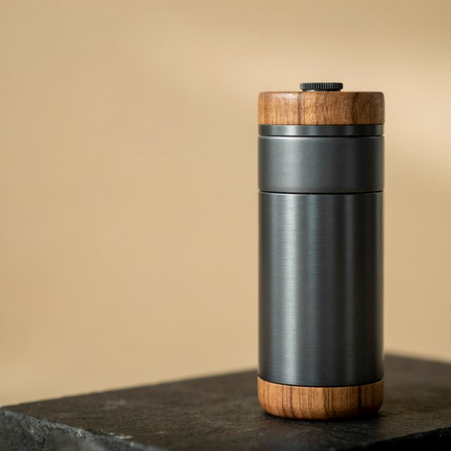 Commercial product photograph of an unbranded portable espresso grinder crafted from anodized aluminum with walnut accents, resting on a black stone surface against a warm beige background, illuminated by broad diffused studio light and a subtle edge highlight. — Ideogram v4.5