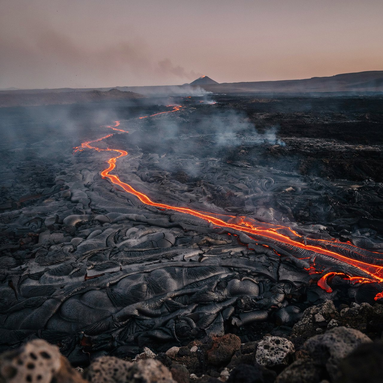 Fresh lava threads a black cinder plain on a volcanic island at dusk beneath ash-laden wind; blistered crust and brittle pumice in the foreground give way to glowing fissures fading into smoke. — Ideogram v4