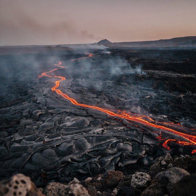 Fresh lava threads a black cinder plain on a volcanic island at dusk beneath ash-laden wind; blistered crust and brittle pumice in the foreground give way to glowing fissures fading into smoke. — Ideogram v4