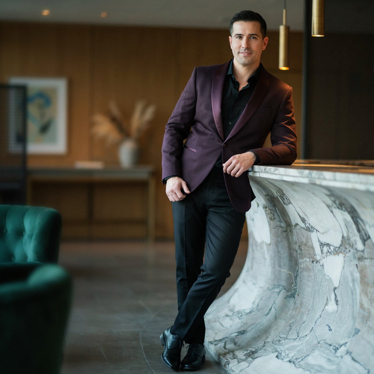 Editorial glamour photograph of an adult man in a midnight-plum dinner jacket and black trousers, standing naturally beside a sculptural marble bar in a quiet hotel lounge, flattering amber light, easy smile, polished editorial elegance. — Ideogram v4