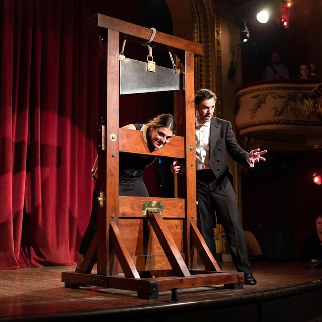 Well dressed stage magician performs the guillotine illusion using a beautiful glamorous adult female assistant. The smiling confident assistant has her head through the hole in the guillotine device. — ImagineArt 2