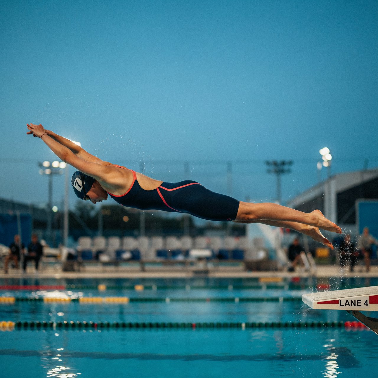 Under the blue-hour floodlights, an adult swimmer drives off the starting block into an outdoor pool. A real-world sports press photo, shot at 200mm with shallow depth of field, captures beads of water suspended around her shoulder. — Ideogram v4