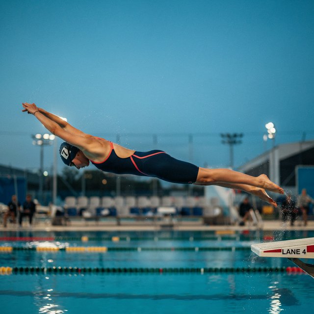 Under the blue-hour floodlights, an adult swimmer drives off the starting block into an outdoor pool. A real-world sports press photo, shot at 200mm with shallow depth of field, captures beads of water suspended around her shoulder. — Ideogram v4