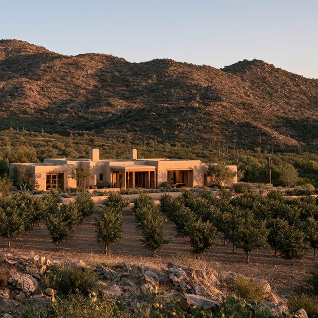 Beyond dry orchard rows, a Pueblo Revival adobe house settles into the desert foothills, with timber lintels and a muted clay roof; warm late-afternoon light, viewed from a rocky rise, reveals clean geometry and credible construction. — Midjourney V8.2