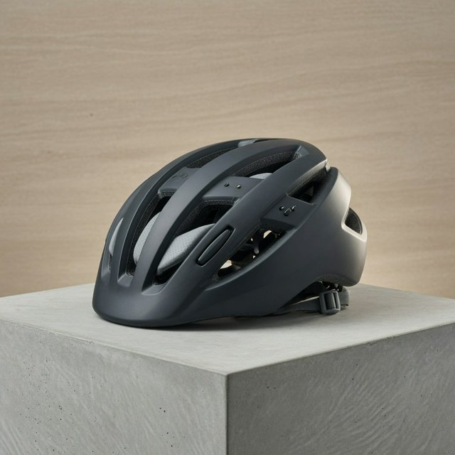 Commercial product photograph of an unbranded folding bicycle helmet made from matte charcoal polymer with soft gray padding, resting on a honed concrete plinth against a muted sand background, illuminated by a broad overhead softbox and gentle rim light. — Grok Imagine Image Quality