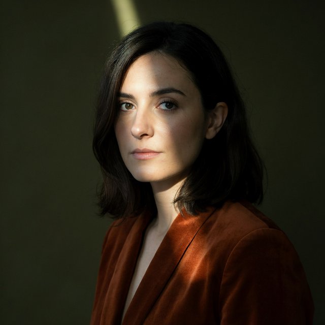 Her guarded gaze catches a solitary beam in a low-key fashion portrait: an adult woman aged 27–35 wears a rust-colored velvet blazer, the rest of her face falling into shadow against a deep olive backdrop. — Ideogram v4.5