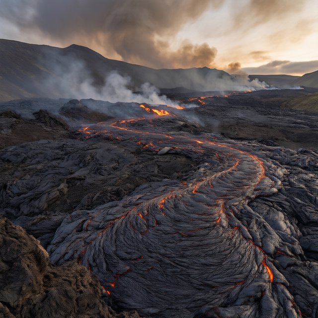 Fresh lava threads a black cinder plain on a volcanic island at dusk beneath ash-laden wind; blistered crust and brittle pumice in the foreground give way to glowing fissures fading into smoke. — Qwen Image 3.0