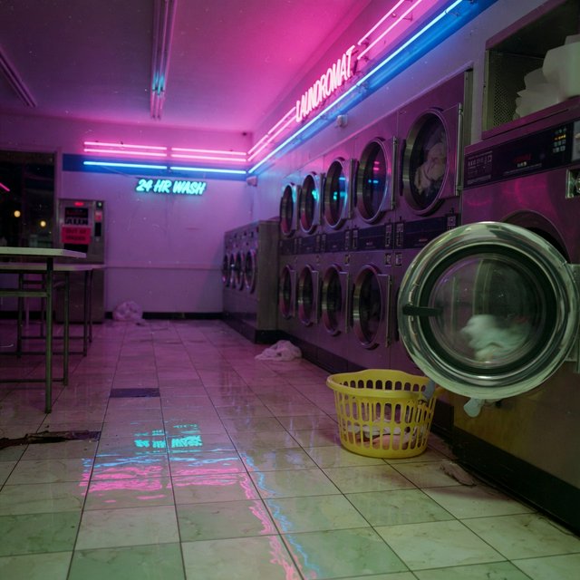 Neon washes a nearly empty overnight laundromat, reflections trembling across tiled floor and spinning steel drums. A handheld street snapshot under magenta and cyan tubes, shot at 35mm with moderate depth of field, catches lint drifting in the air. — Grok Imagine Image Quality