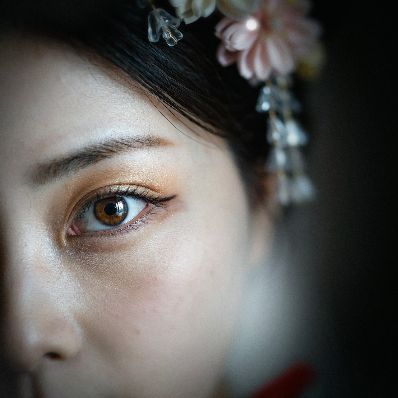 Extreme close-up macro photography of a Geisha's face. The focus is on her eyes, revealing intense emotion and hyper-realistic detail. Her skin is covered in traditional white 'Oshiroi' makeup, showing subtle, realistic cracking and skin texture. Dramatic Chiaroscuro lighting with deep shadows and a single soft light source highlighting the shimmer in her golden eyeshadow and the wetness of her eyes. Ornate kanzashi (hair ornaments) visible in the blurred periphery with intricate silk flowers and dangling silver petals. Cinematic composition, shot on 100mm macro lens, incredibly detailed eyelashes and iris patterns. — Ideogram v4